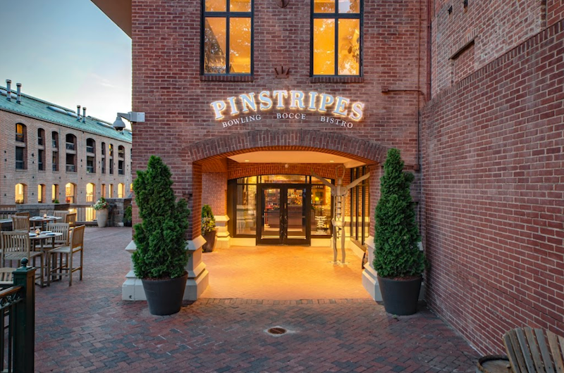 The front exterior of Pinstripes has some greenery in front of a brick building.